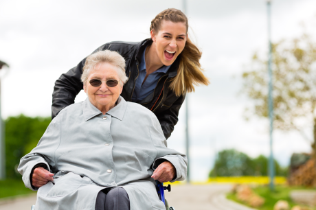 Junge Frau schiebt ältere Frau im Rollstuhl, beide lächeln